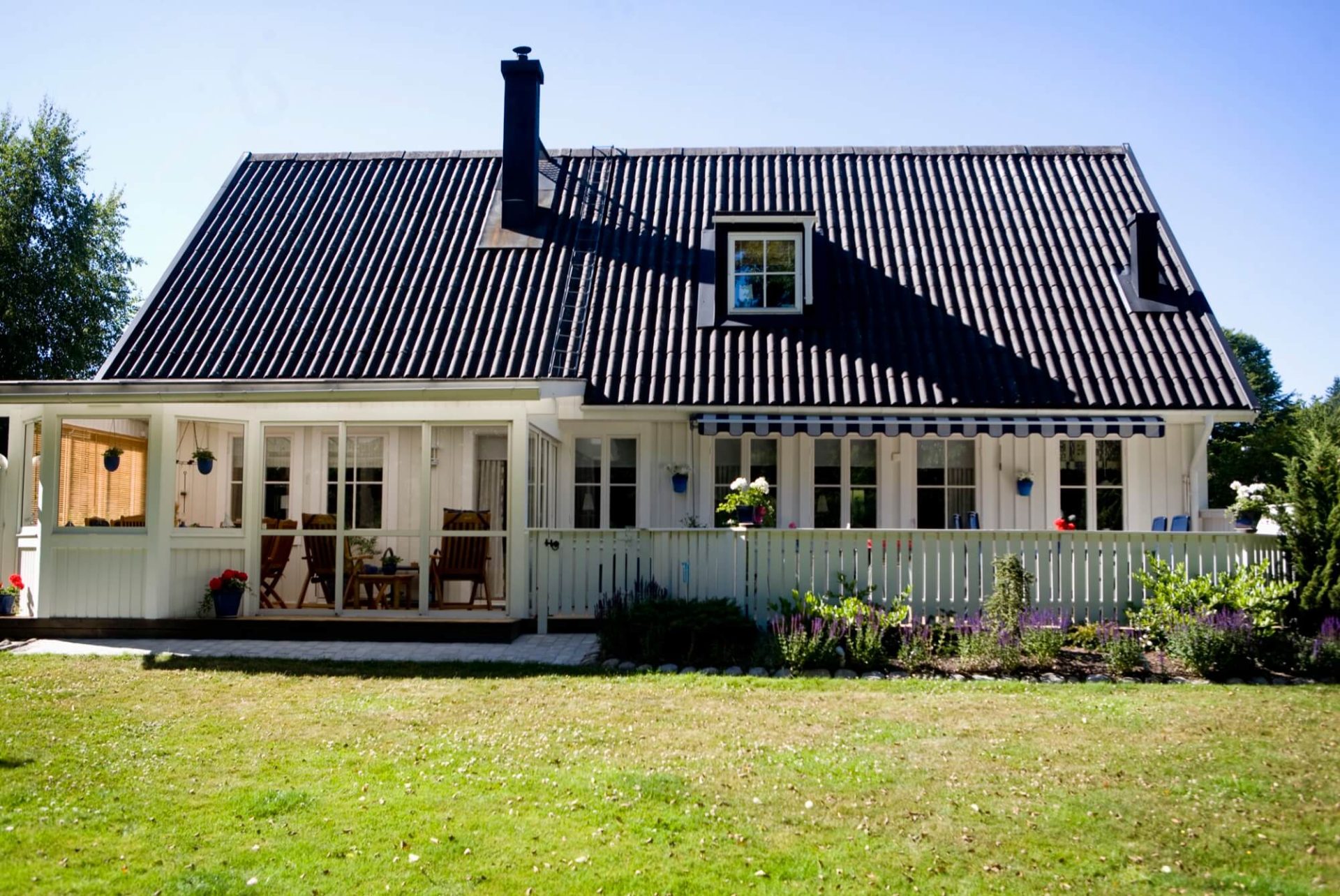 En imponerande vit villa i trä, med ett mörkt tak, omgiven av en generös veranda och en trädgård full av grönska